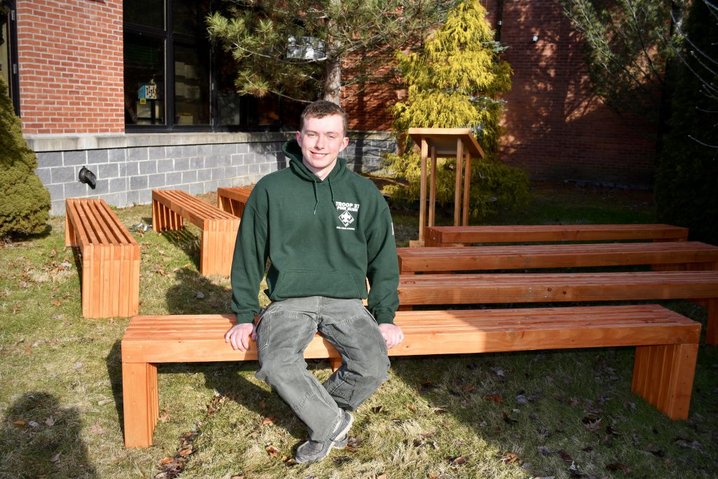 A high school young man in a green hoodie sits on a bench he made. He is smiling.
