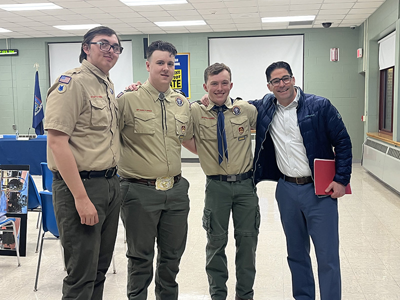 Three high school boys stand with a man, who is wearing a white shirt and dark jacket. The boys are wearing scout uniforms.
