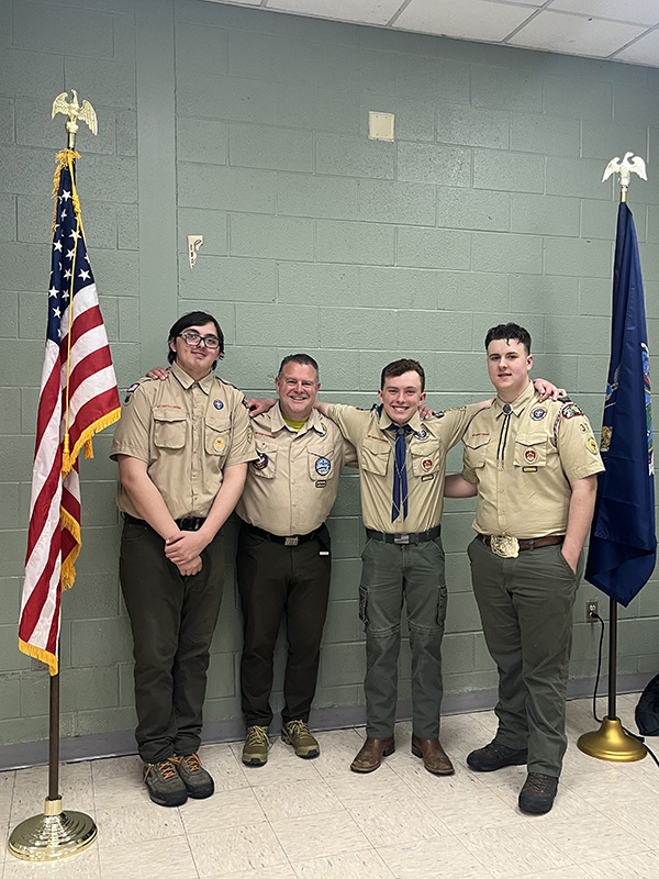 Three high school boys stand with a man on the right. On the left is the American flag. They all are wearing scout uniforms.