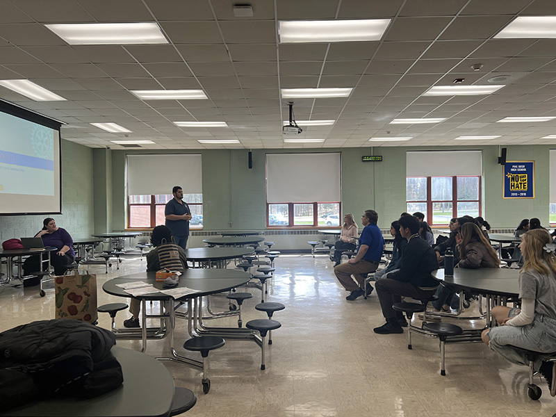 A large room with kids sitting at tables listening to a speaker at the front of the room.