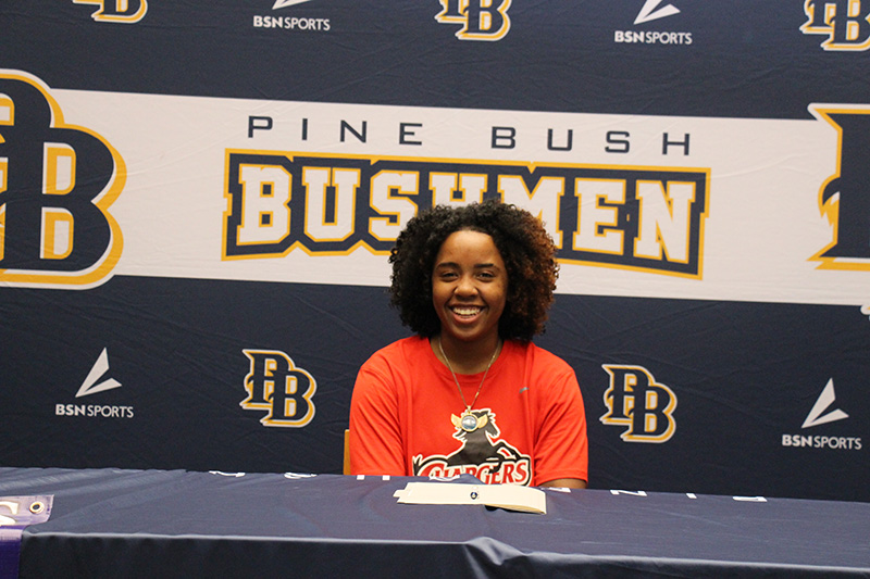 /a young woman in an orange shirt sits at a table with a piece of paper in front of her. She is smiling and the backdrop says Pine Bush Bushmen.