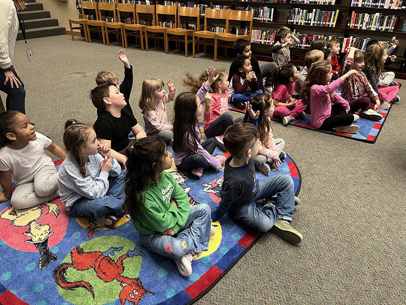 A group of kindergarten students sit on rugs raising their hands.