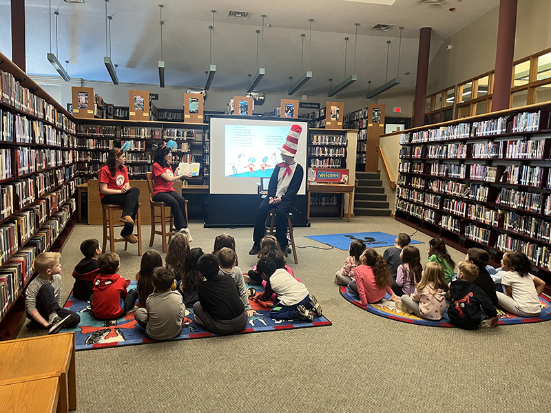 Photo taken from the back - two high school students dressed at Thing 1 and Thing 2 read books to a group of kindergarten students sitting on the floor. There is a high school students dressed as the Cat in the Hat.