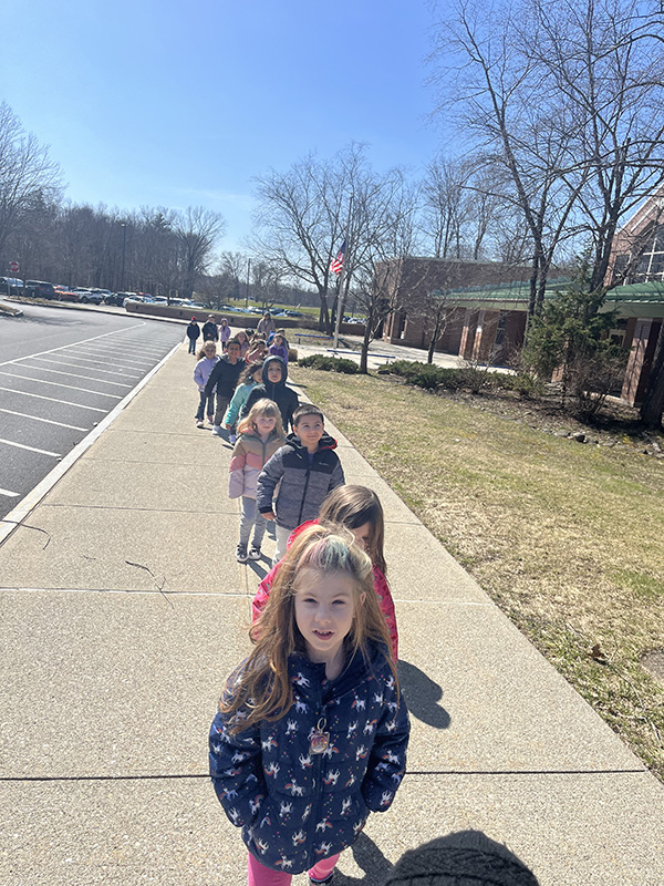 A long single-file line of kindergarten students walking  on a sidewalk. It's a sunny day with bright blue skies.