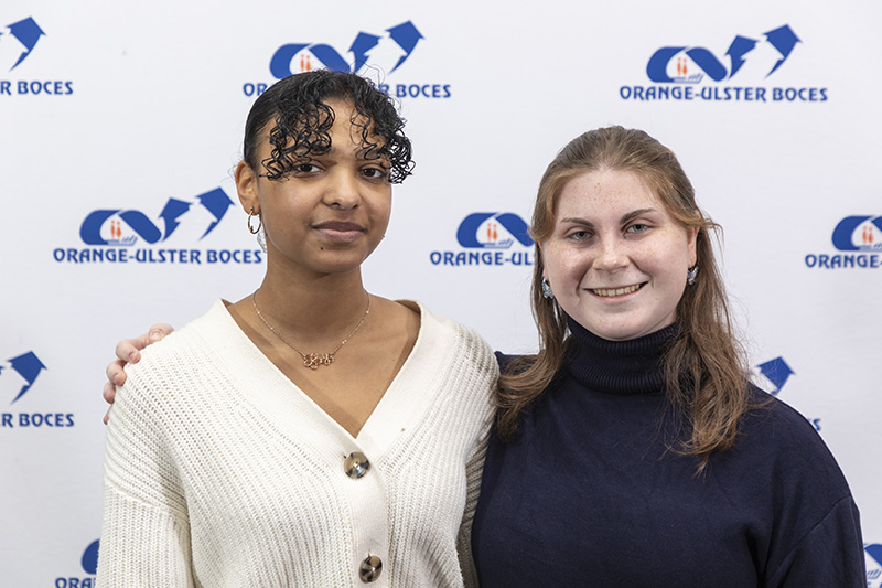 Two high school girls stand arm in arm in front of a blue and white backdrop that says OU BOCES. They are smiling.