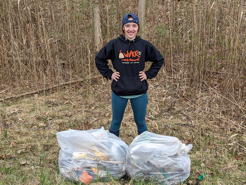 A high school student with a backwards baseball cap, wearing jeans and a black shirt, stands in front of two large white bags of trash.