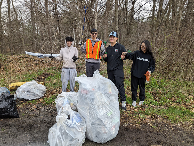 Four high school students, some holding pickers and others holding interesting pieces of trash including a hood ornament for a Subaru, stand by several large bags of trash.