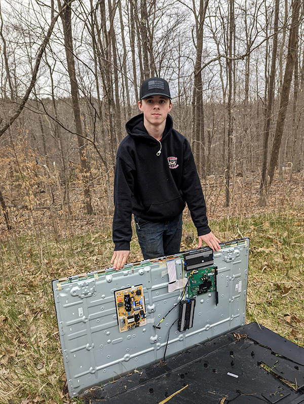 A young man dressed in black, holds a large old television up which he found during a road clean up. 