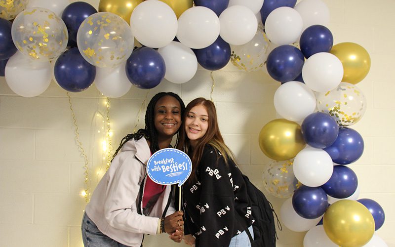 Two girls smile and hold a little sign that says Breakfast with my Bestie. They are under a blue, white and gold balloon arch.