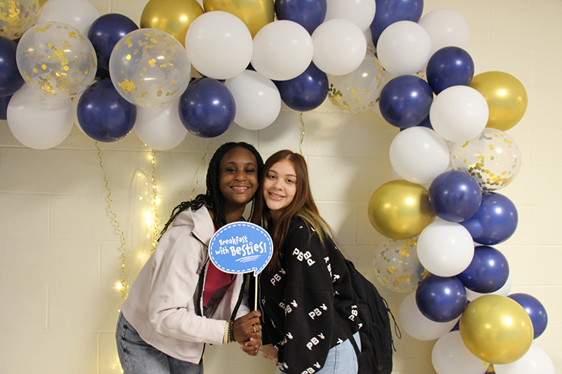 Two girls smile and hold a little sign that says Breakfast with my Bestie. They are under a blue, white and gold balloon arch.