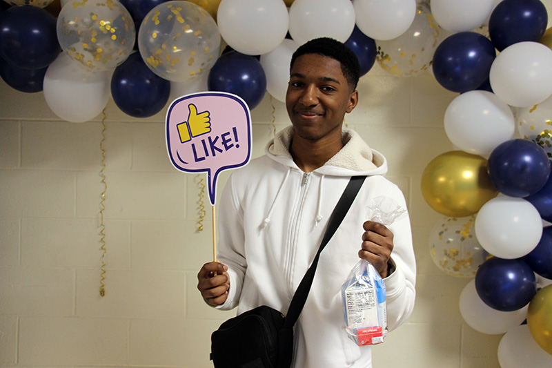 A high school boy wearing a white hoodie holds his bagged breakfast in one hand and a sign that says Like with a thumbs up.