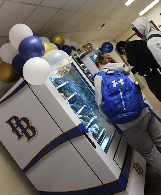 High school kids stand near a vending machine  that has a big blue and gold PB on it.
