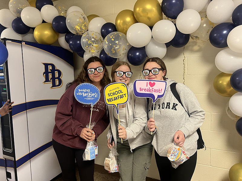 Three girls stand under a gold, blue and white balloon arch. They are holding little signs and smiling, and wearing fun glasses.
