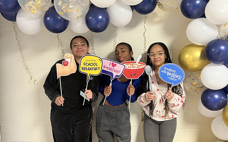 Three high school girls stand under a blue, gold and white balloon arch. They are holding little signs and smiling.