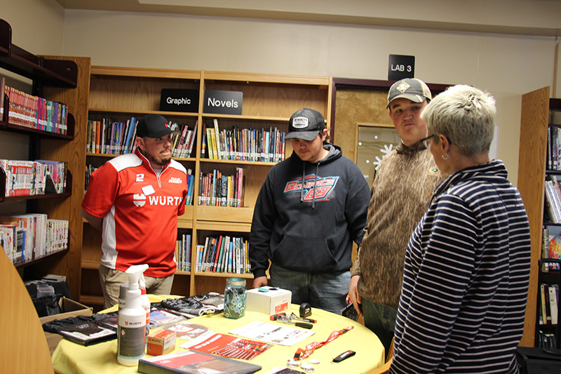 A man is talking to two young men at a table with literature on it. There is a woman standing with them.