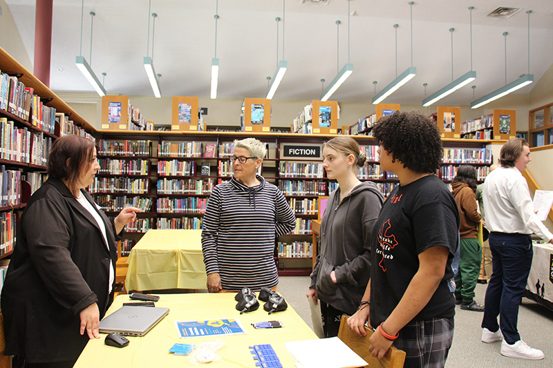 A woman and two high school students stand at a table listening to a woman. They are in a library with books surrounding them.