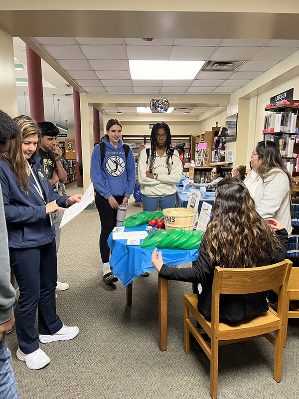 Two women on one side of a table talk to high school kids on the other side. There are green Frisbees on the table for kids to take.