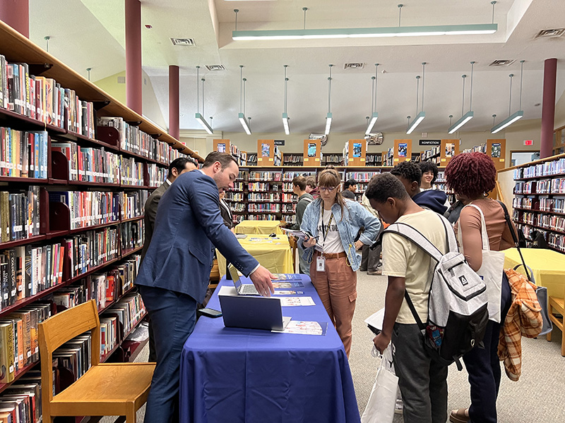 A man points to something on a table as a group of high schools students talk to him.