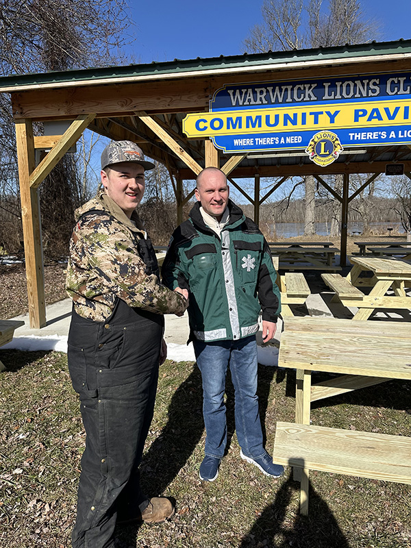 A high school young man wearing a hat and jacket shakes the hand of a man wearing a green jacket. There are picnic benches behind them.