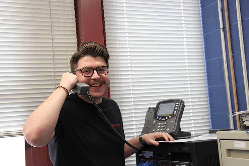 A high school student wearing a black shirt, glasses and with short dark hair, holds a phone and smiles.