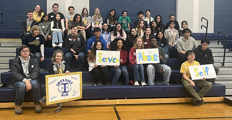 A group of high school and middle school kids sitting on bleachers. There are four rows of them and some are holding signs that say service above self.
