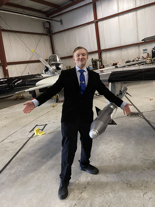 A high school young man wearing a suit and tie, posing in front of an airplane. He is smiling and has his two arms out wide.
