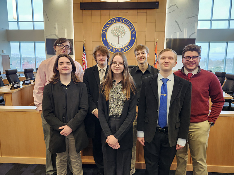 A group of seven high school students stand together , all are dressed nicely. They are in front of a wooden dais with a government seal behind them.