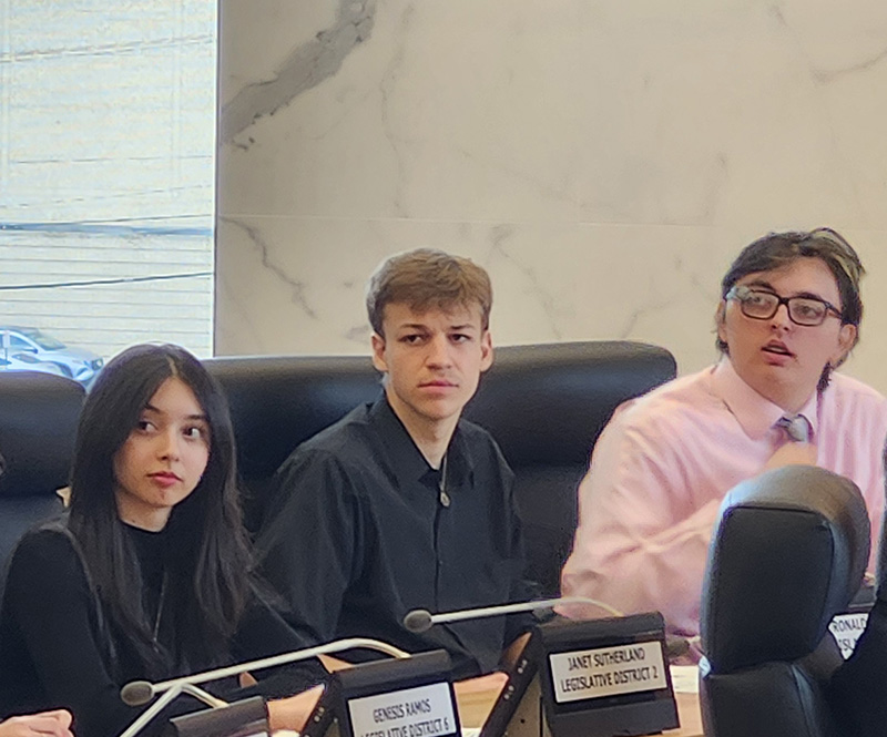 Three high school students sit at desks in a government center.