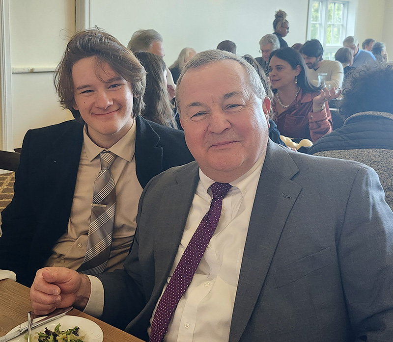 A high school student in a suit and tie sits next to an older man, also in suit and tie. They are smiling.