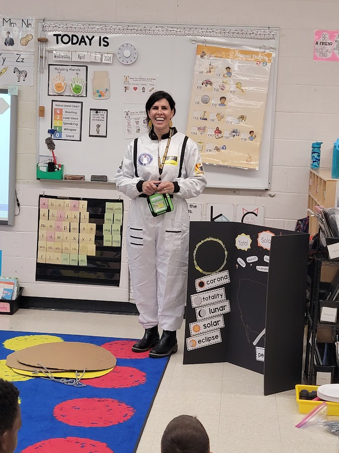 A woman with long dark hair smiles in front of a classroom. She is wearing a one-piece white astronaut outfit.