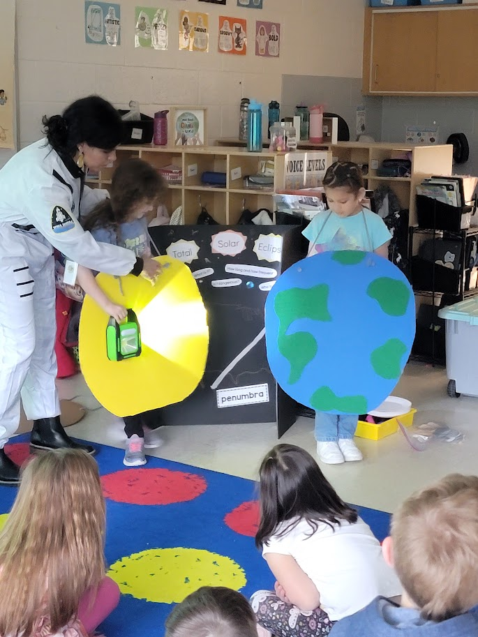 A teacher in a white astronaut costume stands in front of a classroom of students. Some of the elementary students are representing the earth, sun and moon for an eclipse lesson.