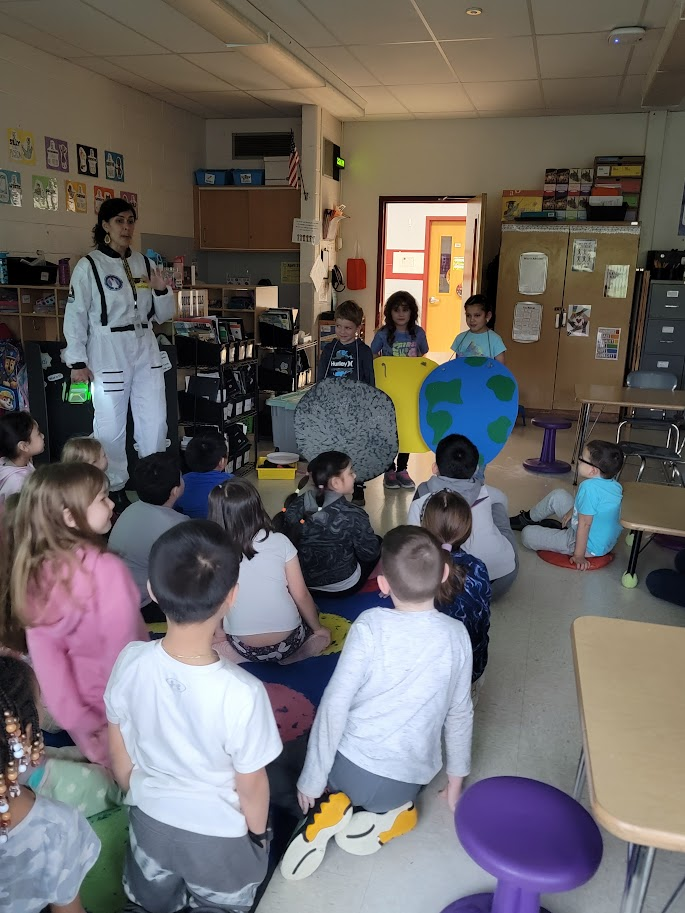 A teacher in a white astronaut costume stands in front of a classroom of students. Some of the elementary students are representing the earth, sun and moon for an eclipse lesson.