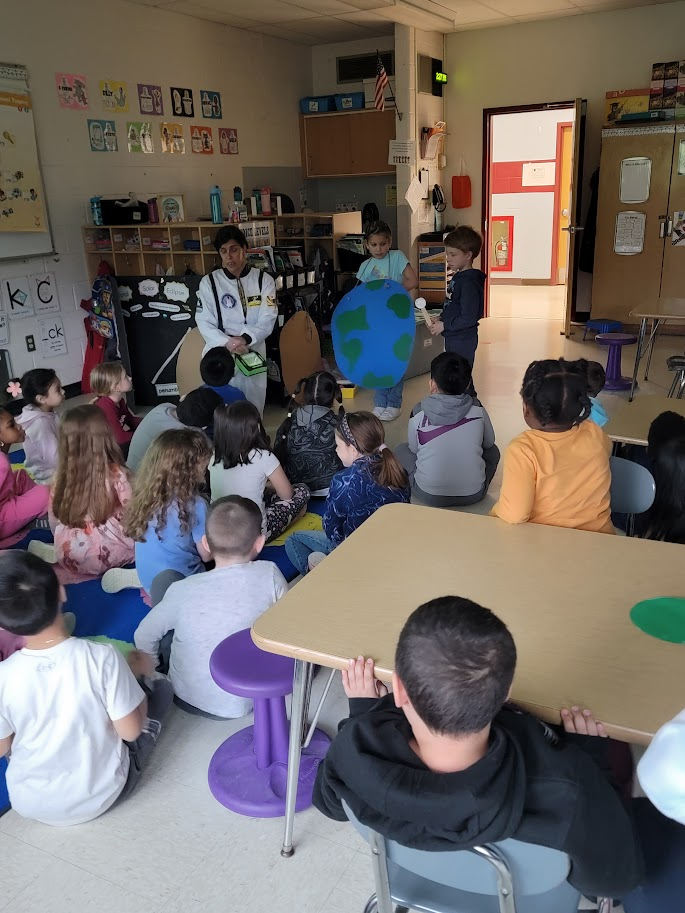 A classroom of first-graders listens to a teacher dressed as an astronaut, while others have a sun, moon or earth placard around their neck. This is a lesson about the eclipse.
