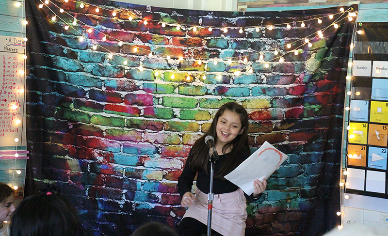 A girl with long dark hair stands in front of fabric that looks like a multi-color brick wall. It has little white lights on it too. She is at a microphone reading poetry.