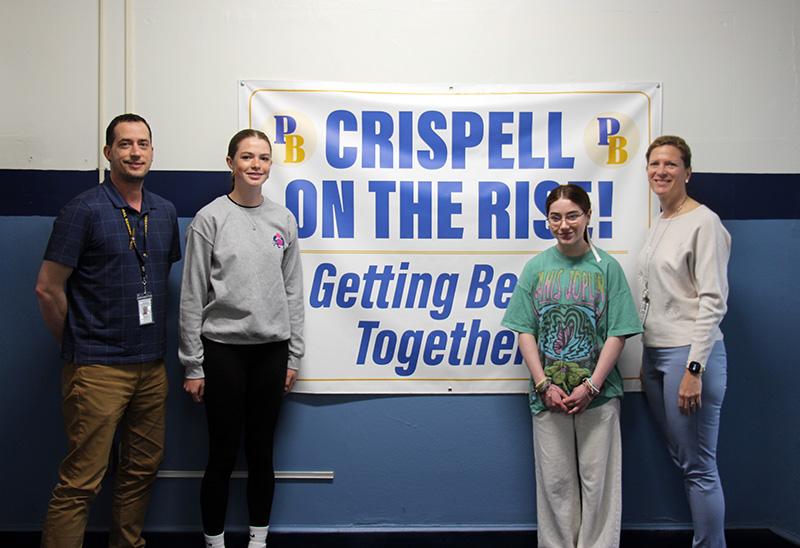 A sign in the center says Crispell on the Rise Getting Better Together. There is a man on the left and an eighth-grade girl with long hair pulled back standing next to him. On the right side, is a woman on the far right and another eighth-grade girl with dark hair pulled back. All are smiling.