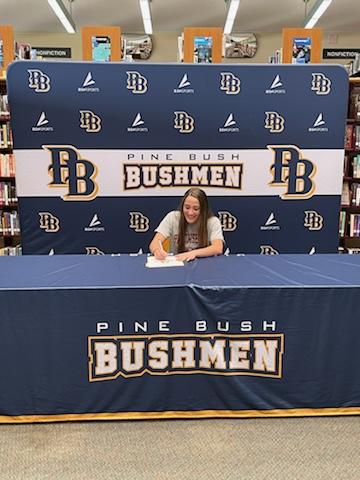 A high school senior sits at a table with a tablecloth that says Pine Bush Bushmen. She is signing a piece of paper.