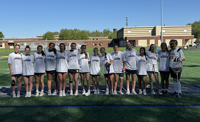 15 high school girls stand together in a straight line. They are wearin gwhite tshirts that say Pine Bush Lacrosse