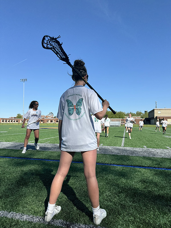 A blue sky and green grass. A high school girl stands with her lacrosse stick over her shoulder. She is wearing a white tshirt with a teal butterfly on it. It says Morgan's Message Taking a shot at mental health.