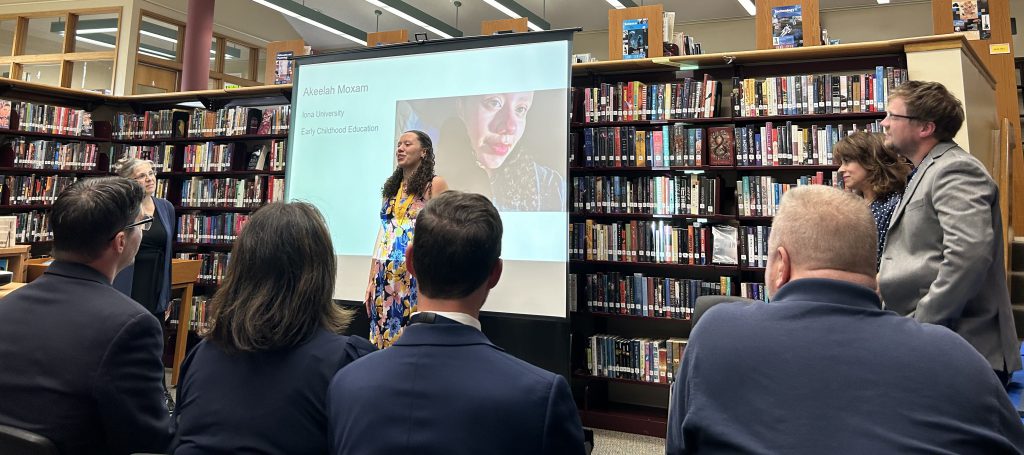 A high school young woman stands in front of a group of people presenting. Behind her is a screen with a picture and information on it.