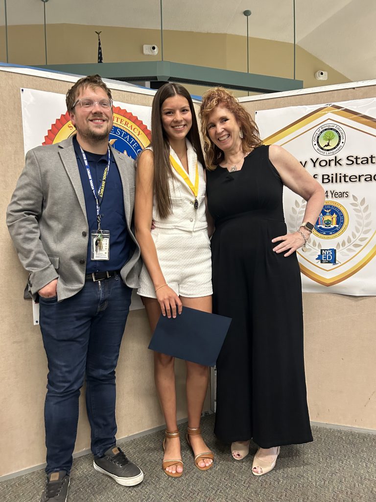 Two adults, a man on the left and a woman in a long black dress on the right, stand by a young woman with long dark hair with a gold medallion around her neck.
