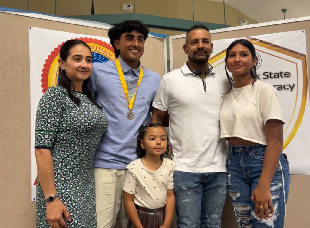 A high school boy, along with his family, two adults, a young woman and a small child, stand together.
