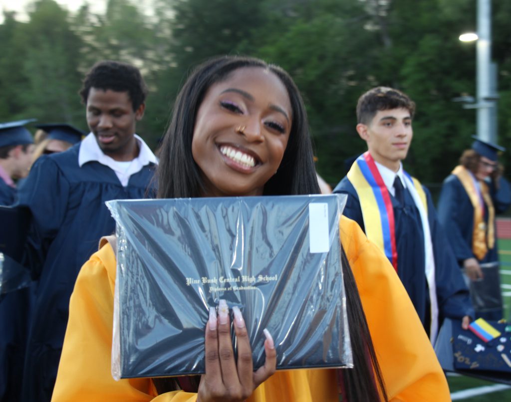 A young woman wearing a gold graduation gown smiles and holds up her diploma.