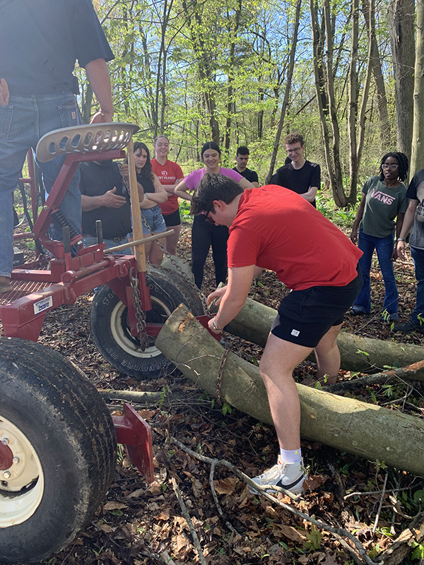 A high school student in black shorts and a red shirt hooks something onto a tractor that is pulling a large log.
