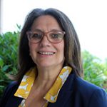 A woman with shoulder-length hair and glasses smiles. she is wearing a yellow and white shirt and navy blue blazer.