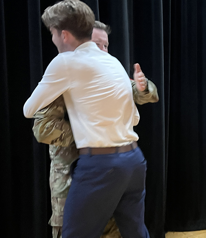 A high school kid in a white shirt and blue pants hugs a man in army fatigues.