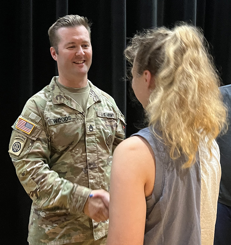 A high school girl with long blonde hair shakes hands with a man in army fatigues. He is smiling.