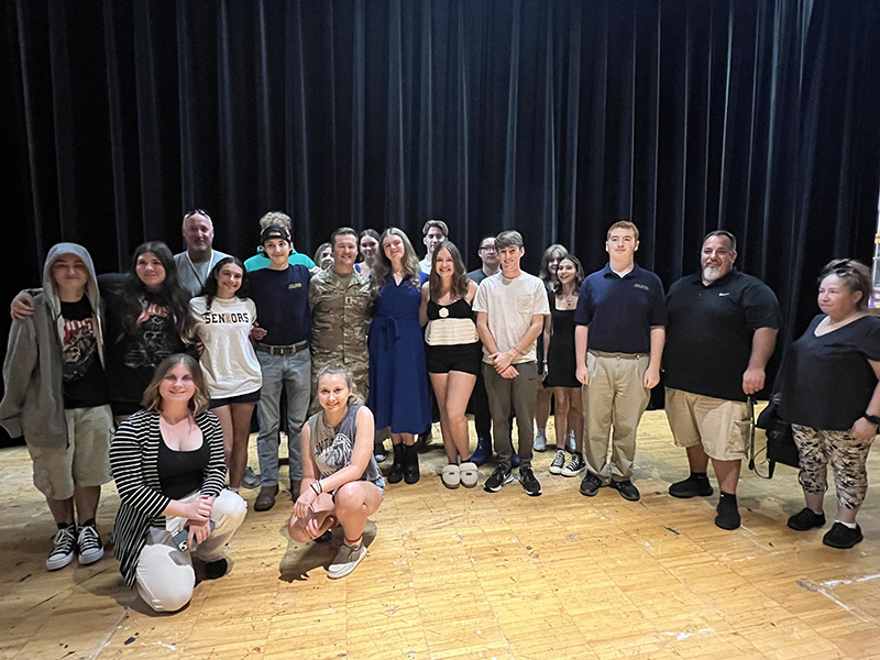A large group of high school kids pose with a man in the center who is wearing army fatigues.