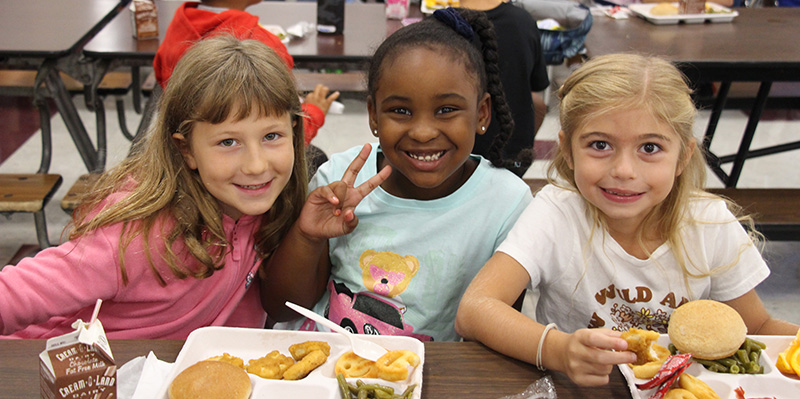 Three girls sit at a table with lunch trays in front of them. They are all smiling and the girl in the center is giving a peace sign.