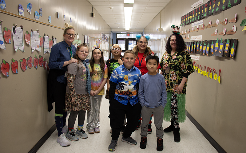 Three adults and five older elementar kids stand together in a hallway. They are all dressed for wacky Wednesday with different colors on, funny prints, head bands.