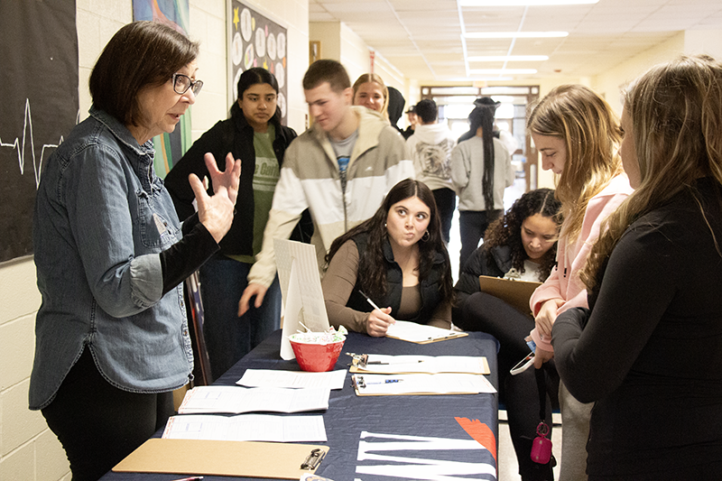 A group of high school kids sit and stand around a long table with a woman on the other side.
